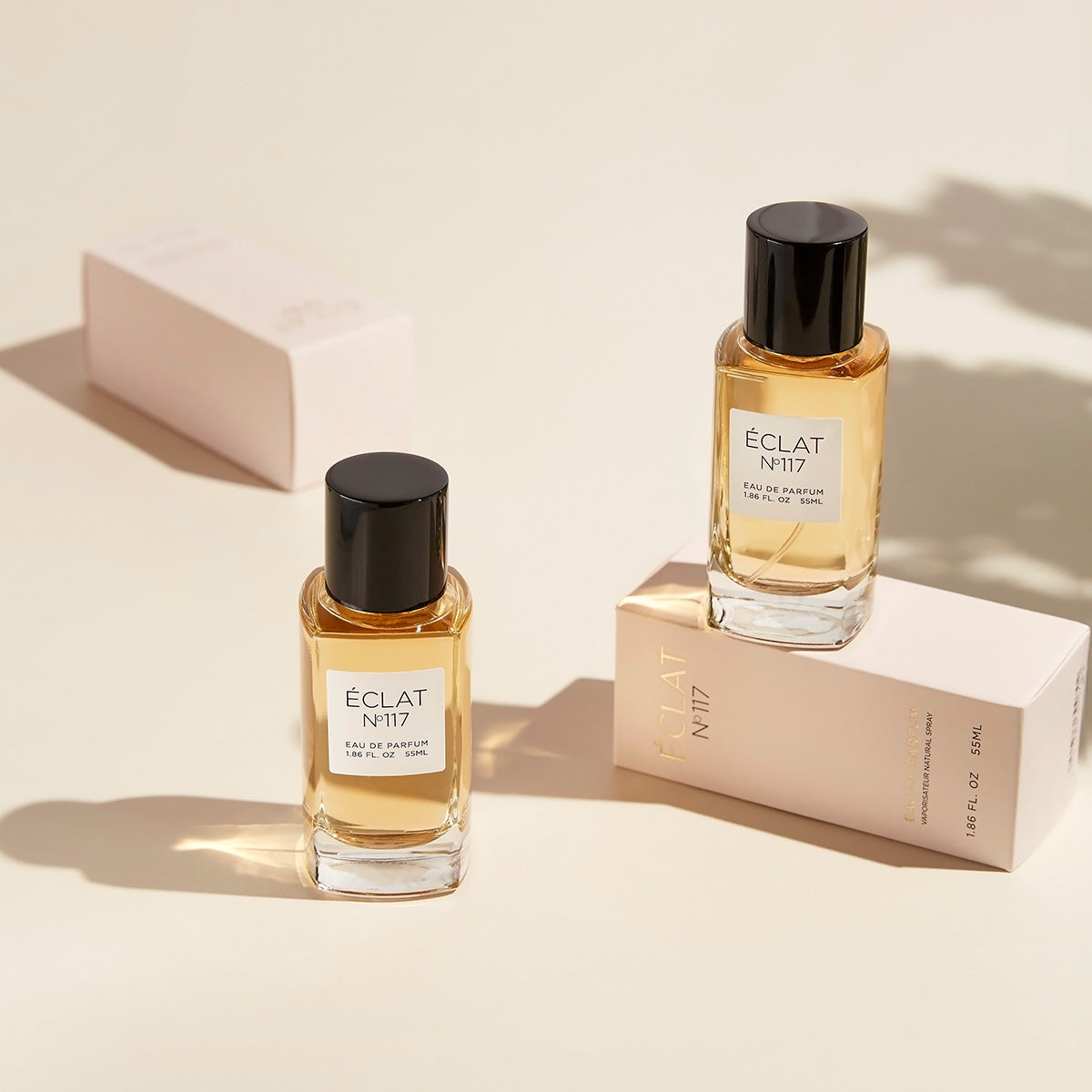 Two bottles of ÉCLAT No. 117 women's perfume displayed on a soft beige surface with matching packaging. The golden liquid in the glass flacons is illuminated by natural light, casting soft shadows for a warm and elegant composition.