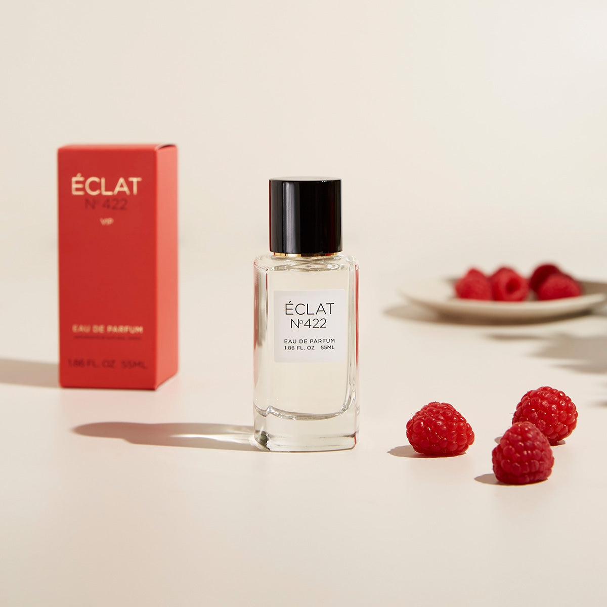 ÉCLAT No. 422 women's perfume bottle placed next to its vibrant raspberry-red VIP packaging, surrounded by fresh raspberries on a cream-colored surface. A small plate with raspberries is blurred in the background, creating a fruity and elegant aesthetic.