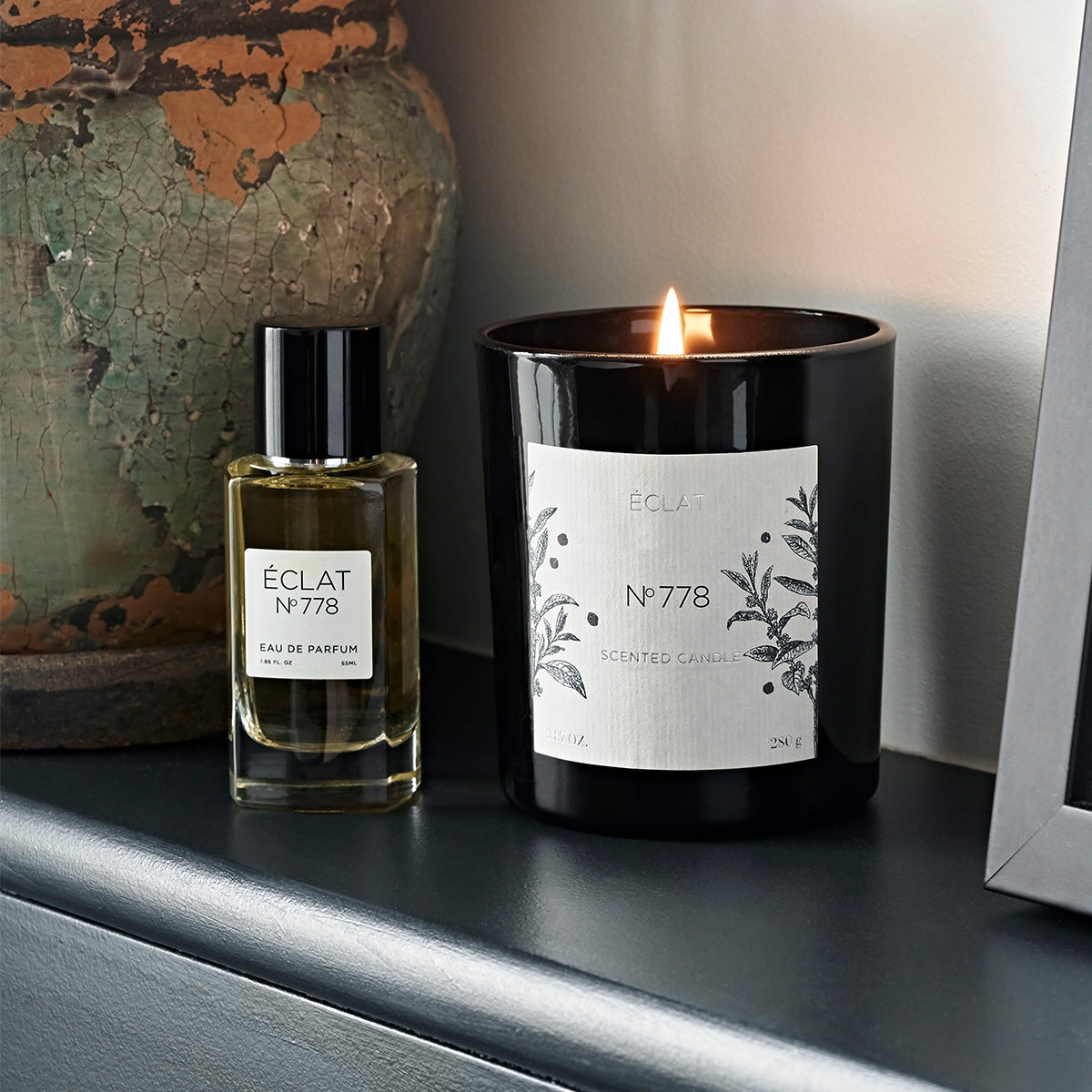 ÉCLAT No. 778 Eau de Parfum and matching scented candle placed on a dark shelf. The perfume bottle has a warm amber hue, and the candle is lit, creating a cozy ambiance. Both items feature minimalist labels with botanical illustrations.