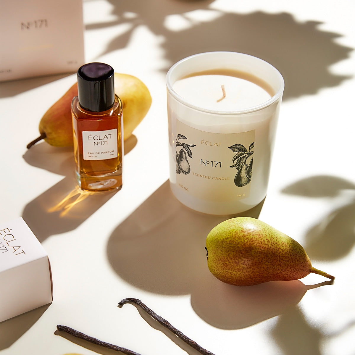 Szklana butelka perfum ÉCLAT Blackcurrant & Praline z czarną nakrętką obok eleganckiej białej świecy i gruszki na jasnym tle.