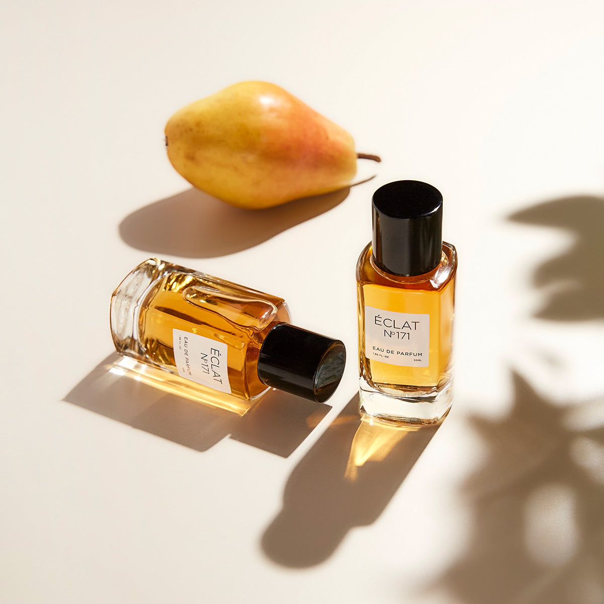 ÉCLAT Blackcurrant & Praline w eleganckich szklanych flakonach z czarnymi nakrętkami, obok dojrzałej gruszki, rzucających miękkie cienie.