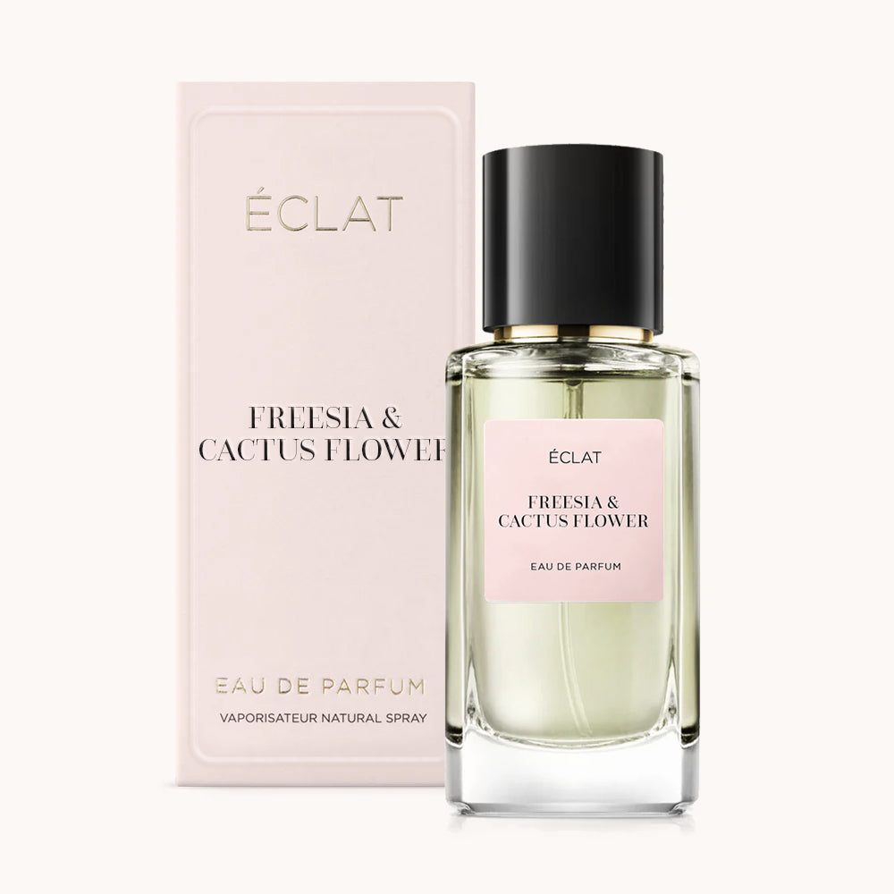 Perfumy ÉCLAT Freesia & Cactus Flower w fasetowanej szklanej butelce z czarną nakrętką, obok eleganckiego bladoróżowego opakowania.
