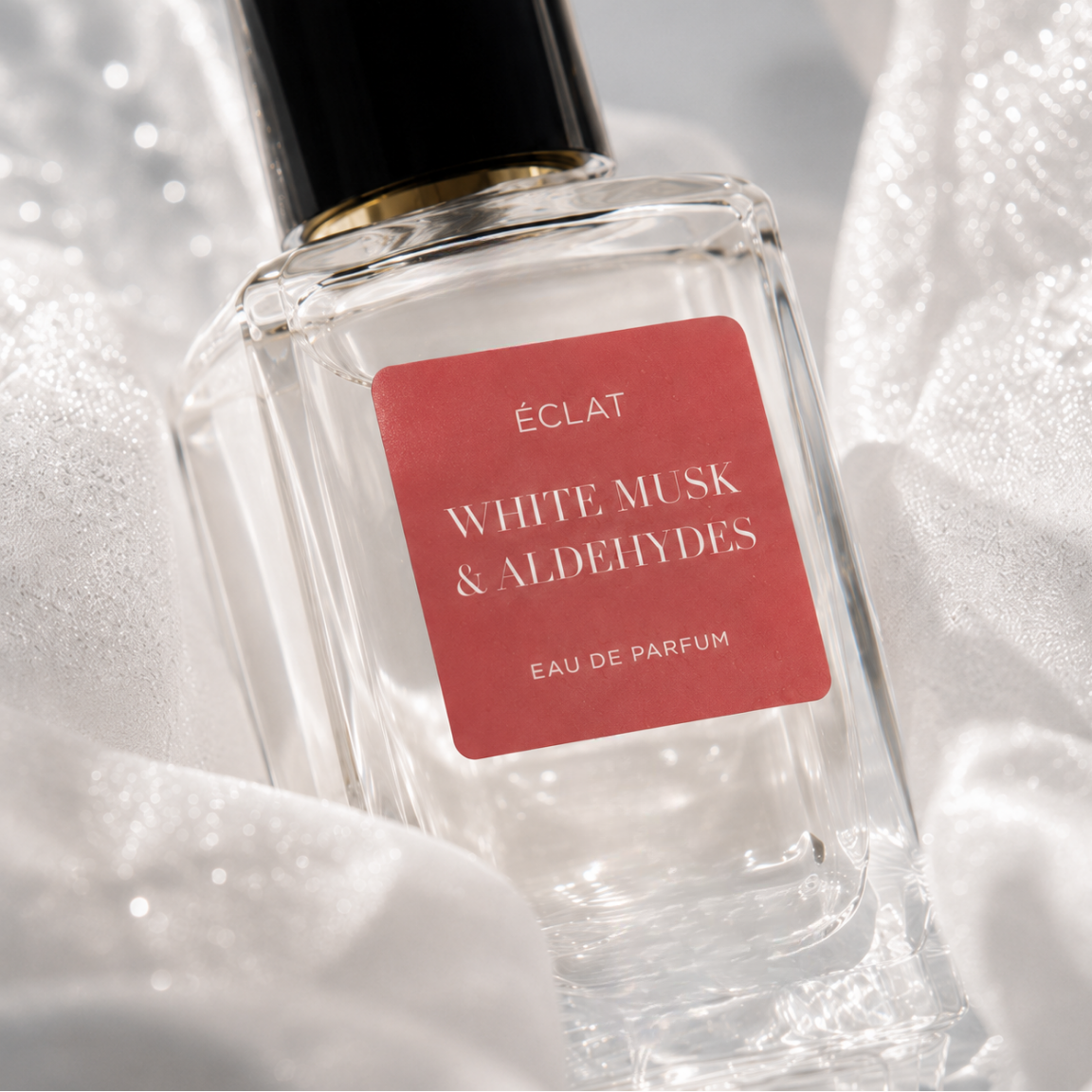 Fasetowana szklana butelka ÉCLAT White Musk & Aldehydes z czerwoną etykietą i czarną nakrętką, otoczona delikatną białą tkaniną.