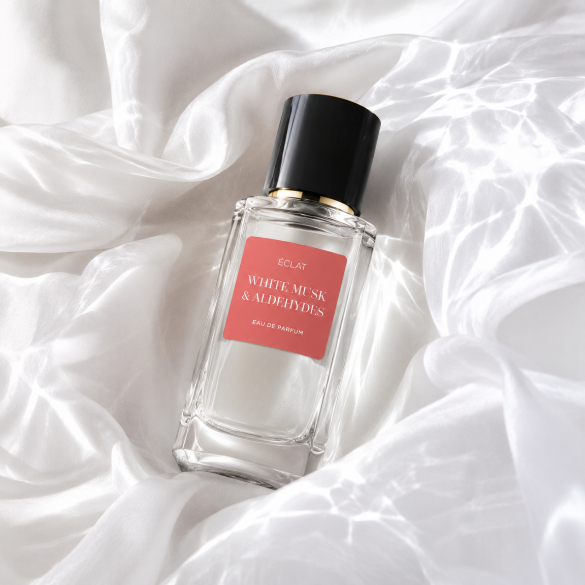 Fasetowany flakon ÉCLAT White Musk & Aldehydes z czarną nakrętką i koralową etykietą na satynowym tle.