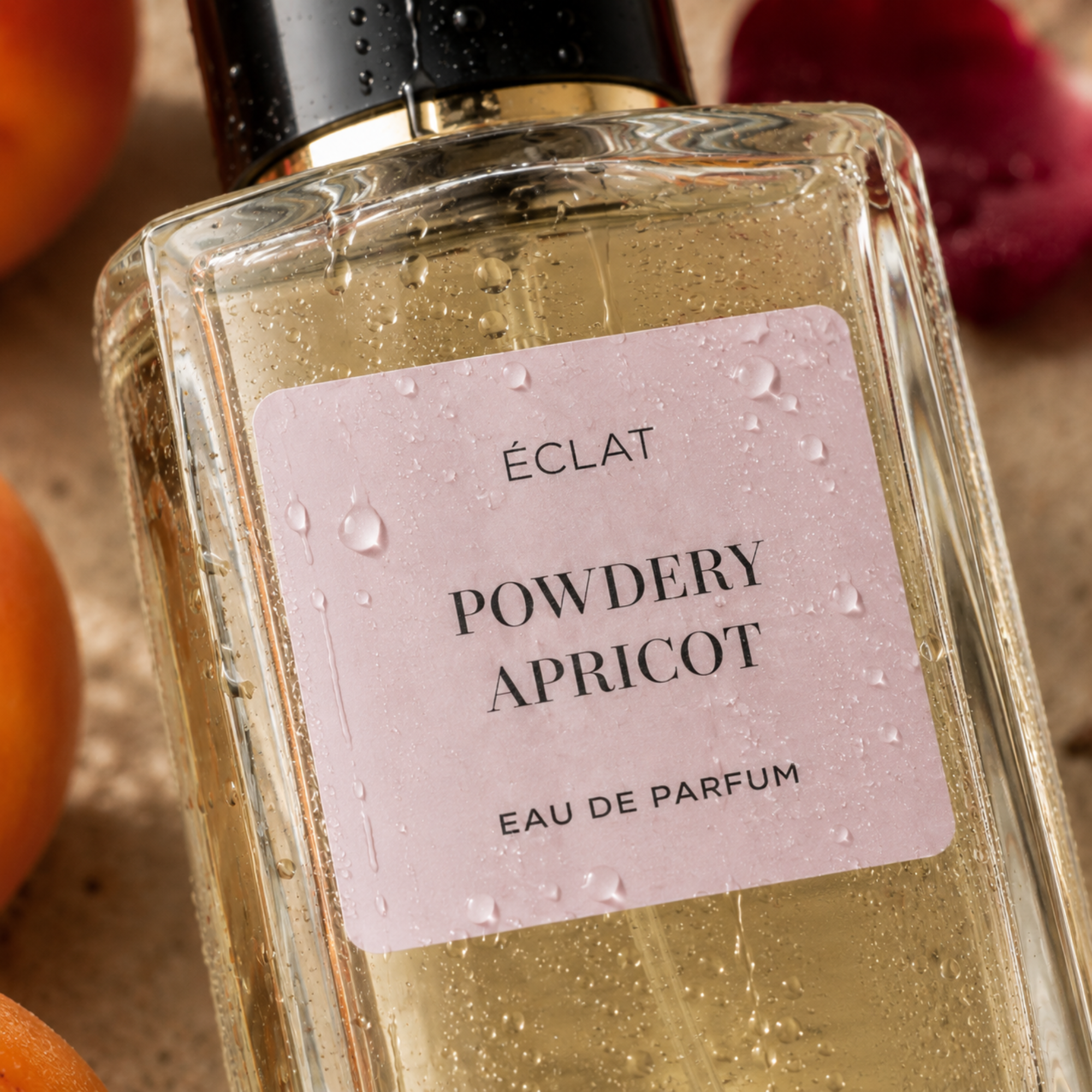 Budka szklana ÉCLAT Powdery Apricot z jasno różową etykietą i czarną nakrętką, otoczona morelą i czerwonymi płatkami.