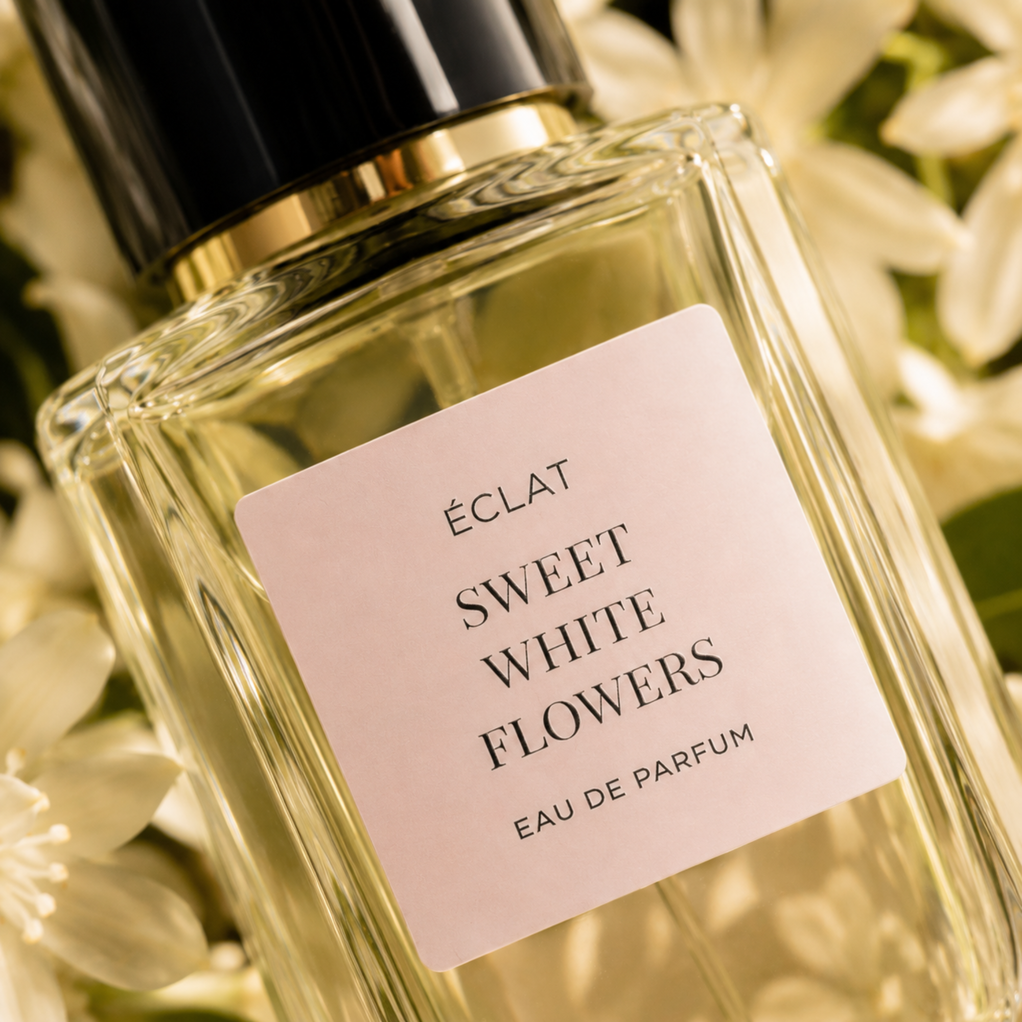 Fasetowana szklana butelka ÉCLAT Sweet White Flowers z delikatnie różową etykietą i czarno-złotą nasadką, otoczona białymi kwiatami.