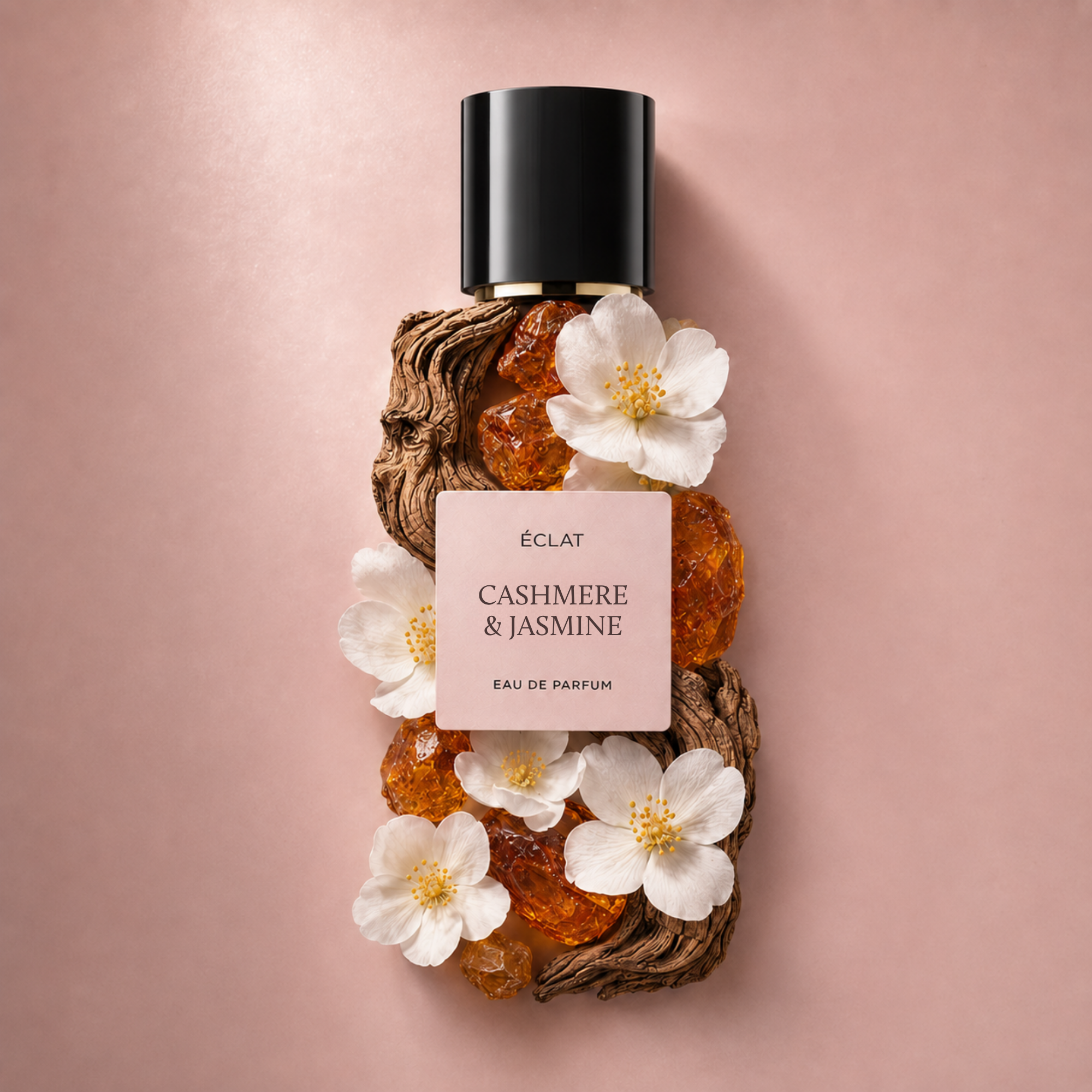 Flakon perfum ÉCLAT Cashmere & Jasmine ozdobiony białymi kwiatami i bursztynowymi kryształami na różowym tle.