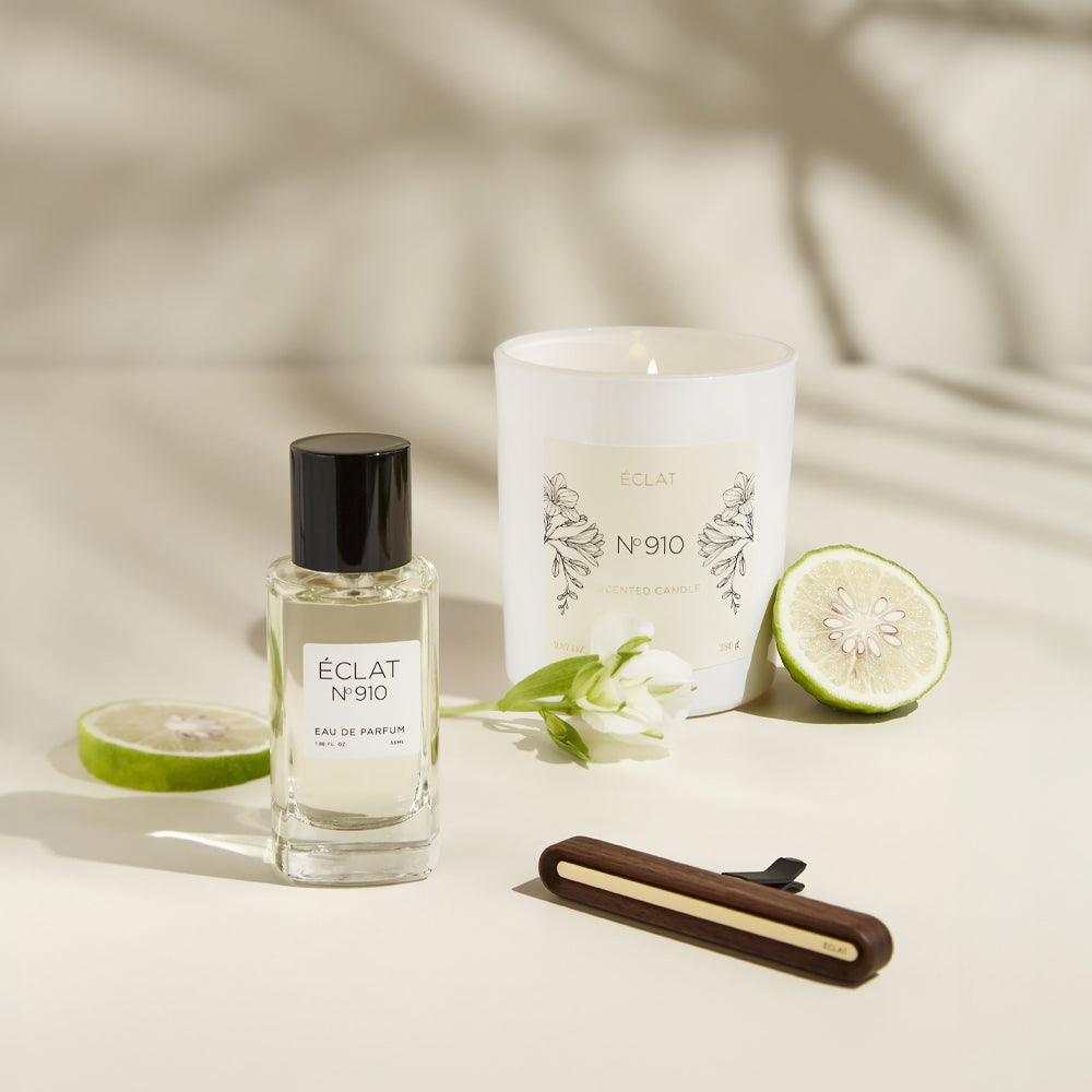Fasetowana szklana butelka perfum ÉCLAT Creamy White z czarną zakrętką, obok białej świecy zapachowej, plaster cytrusa i kwiat.