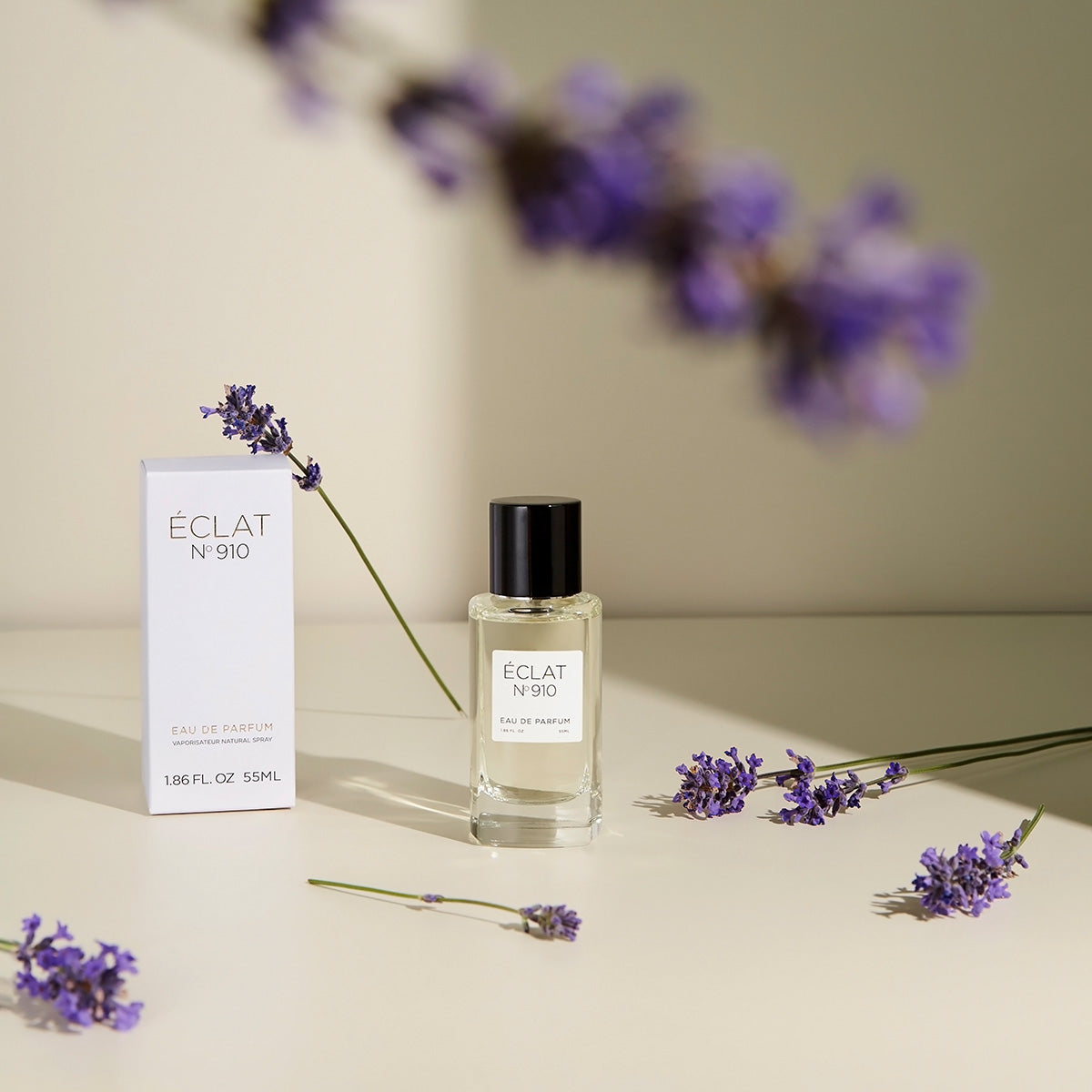 Perfumy ÉCLAT Creamy White w przezroczystej szklanej butelce z czarną nakrętką, otoczone lawendą, obok eleganckiego białego pudełka.