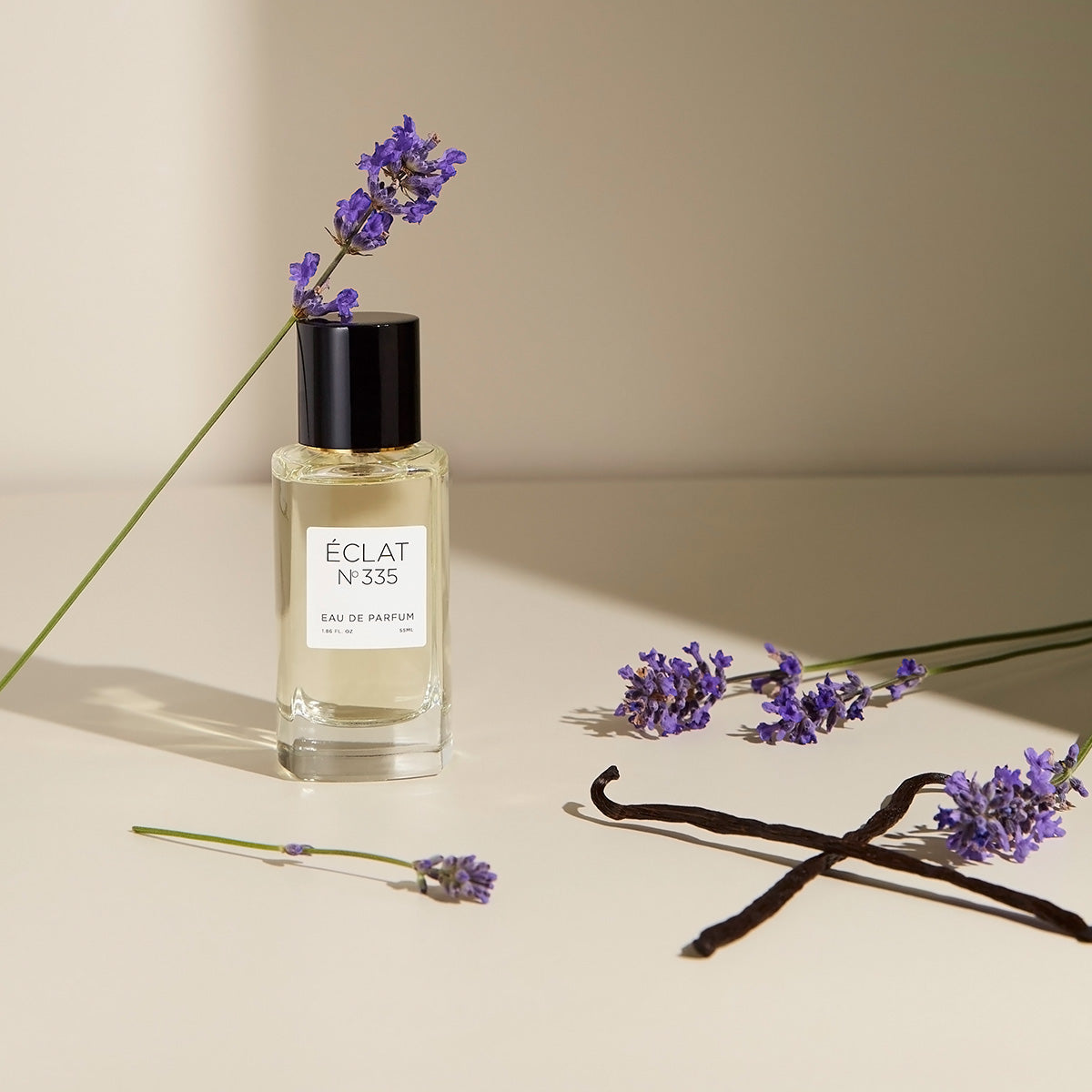 Fasetowana szklana butelka ÉCLAT Lavender & Vanilla z czarną zakrętką, otoczona gałązkami lawendy i strąkami wanilii.