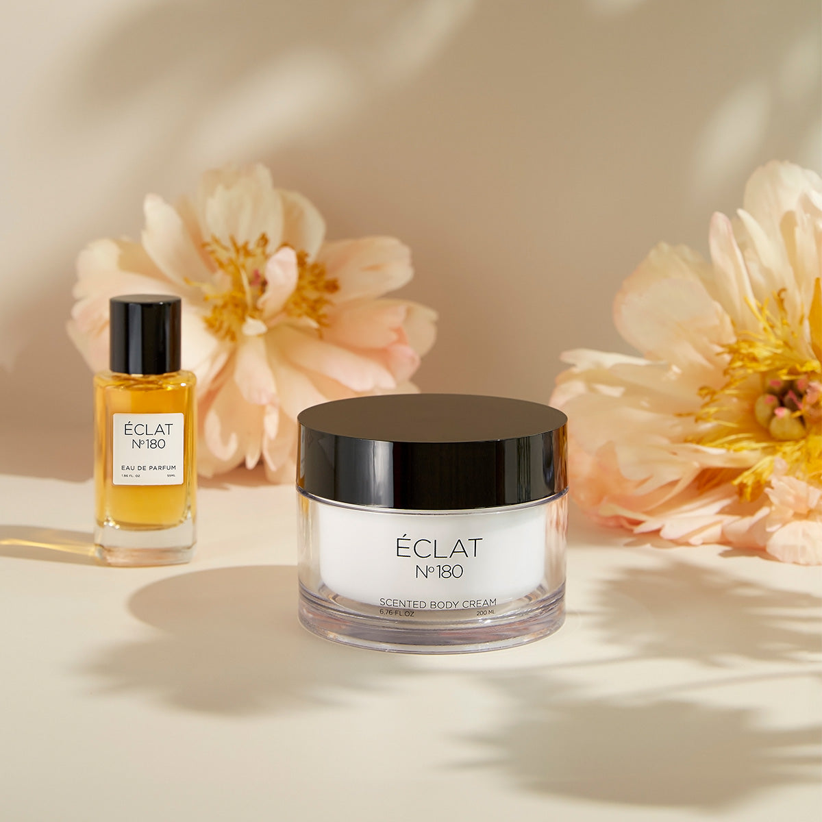 Zestaw ÉCLAT Orchid & Patchouli zawiera flakon perfum z przezroczystego szkła i słoik kremu z czarną pokrywką, otoczony delikatnymi kwiatami.