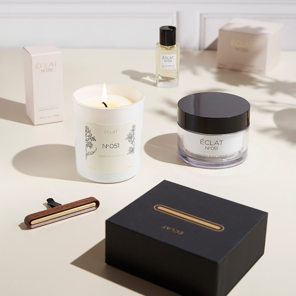 Elegancka kompozycja z zapaloną świecą zapachową, fasetowanym szklanym słoikiem z ciemną pokrywką, butelką perfum i wyrafinowanym pudełkiem, ucieleśniająca ÉCLAT Oriental Almond.