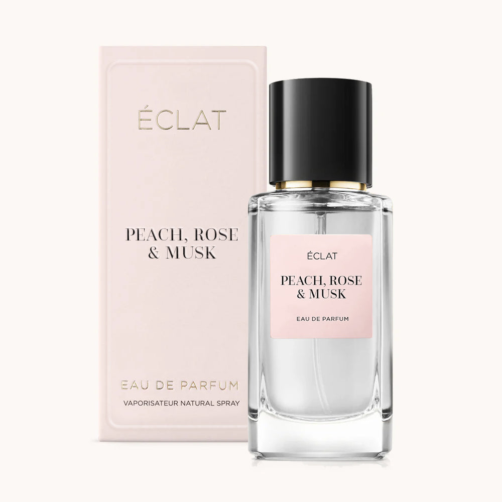 Flakon perfum ÉCLAT Peach, Rose & Musk z przezroczystego szkła z czarną nakrętką, w towarzystwie jasnoróżowego pudełka.