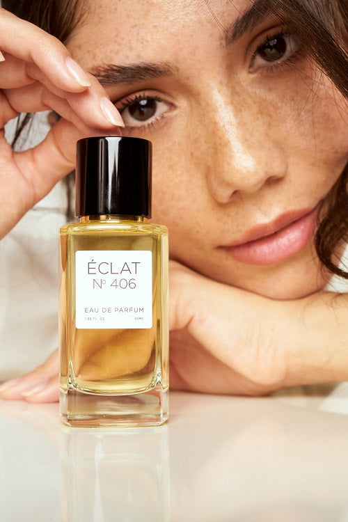 Fasetowana szklana butelka ÉCLAT N° 406 Eau de Parfum z czarną nakrętką przed twarzą osoby.