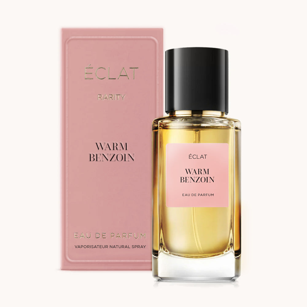 Perfumy ÉCLAT Warm Benzoin są prezentowane w przezroczystym szklanym flakonie z czarną zakrętką i różową etykietą; obok znajduje się pasujące różowe pudełko.